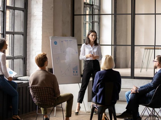 Young confident ceo make forecast financial report on flip chart for diverse staff, group of workmates listen woman business trainer take part at educational meeting in modern office coaching concept
