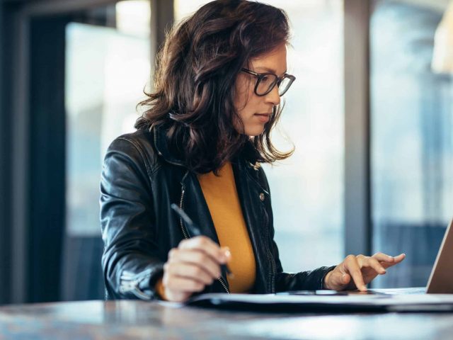 Businesswoman making notes looking at a laptop computer at office. Woman entrepreneur sitting at the table writing notes while working on laptop.